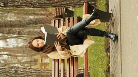 Young attractive female sitting on a bench in park alone reading a book wearing brown trench and scarf. Studying concept. Female reading book in autumn city park. Vertical video
