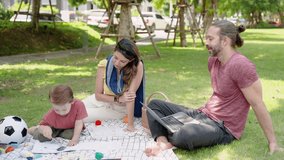 Caucasian family of father, mother and son resting on grass in village park while father works on laptop. male and female couple sit and talk with their children sitting next. boy drawing and coloring