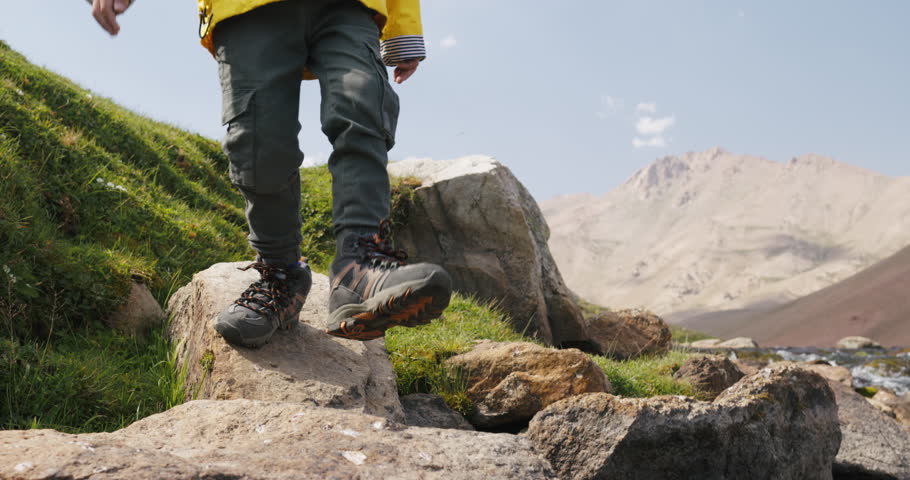 Legs of child hiker crosses stream. Young tourist hiker traveler boy child kid on rocks carefully steps over river on stones. Child boy tourist feet legs stepping over rocks over river stream.