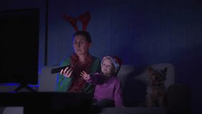 Mom with a little daughter and a small dog are sitting on the sofa near the TV and waiting for the Christmas holiday.New Year and Christmas holiday concept. High quality 4k footage