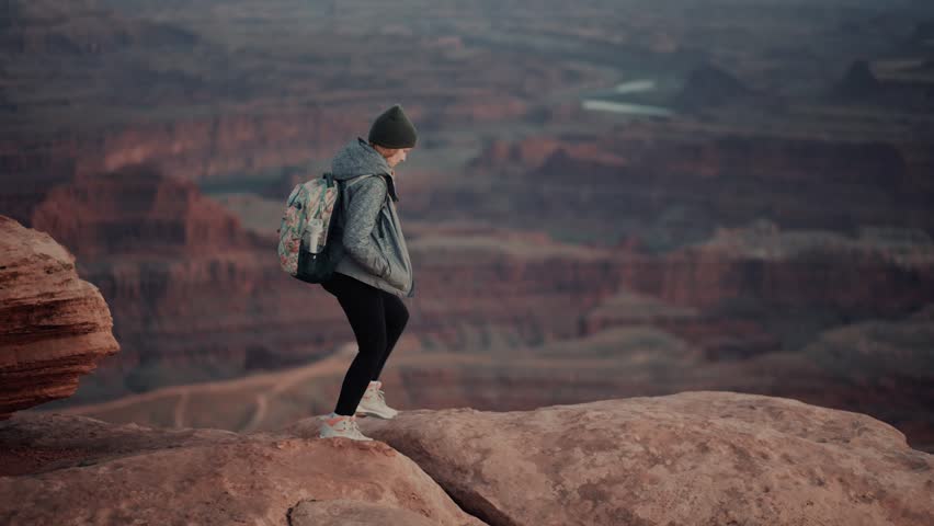 Female hiker walks to the edge of a cliff and admires stunning canyon views during sunset