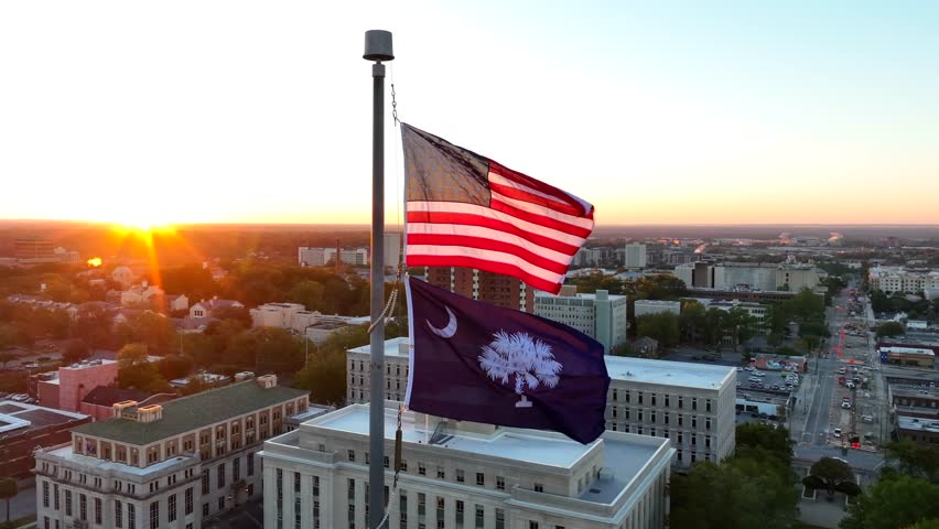 American and South Carolina flags waving atop capitol building in downtown Columbia, SC. Aerial orbit shot with government buildings during bright sunrise.
