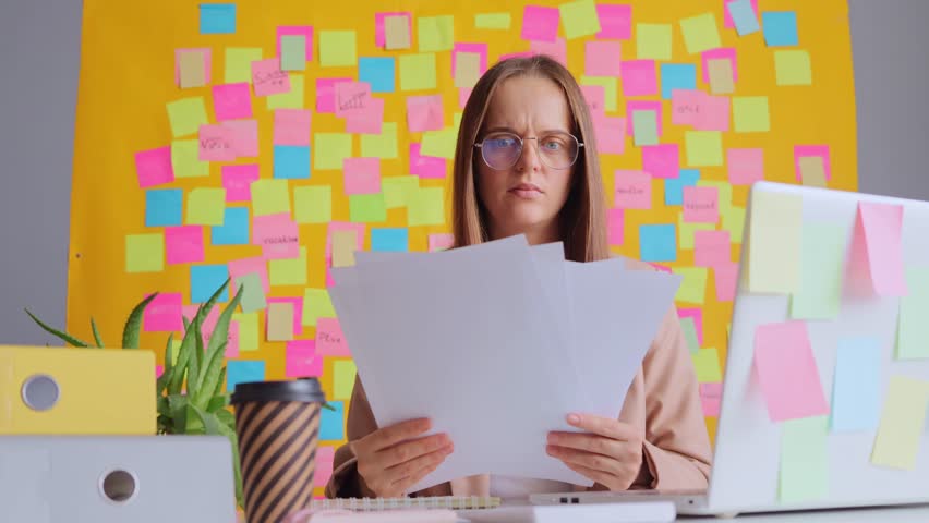 Shocked woman working on computer against yellow wall with colorful stickers finding lots mistakes in her report trying to concentrate on her work.