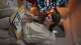 Vertical video. A woman, having painted a Christmas wreath on a postcard in watercolor, positively evaluates her creativity while sitting in a rocking chair against the backdrop of a Christmas tree