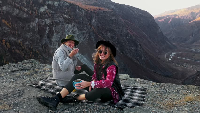 Two traveler female friends sitting on the edge of a sheer cliff above a deep cliff. Girld smilling and lookinh at the camera. Females watching the beauty of nature mountain forest and sunset on