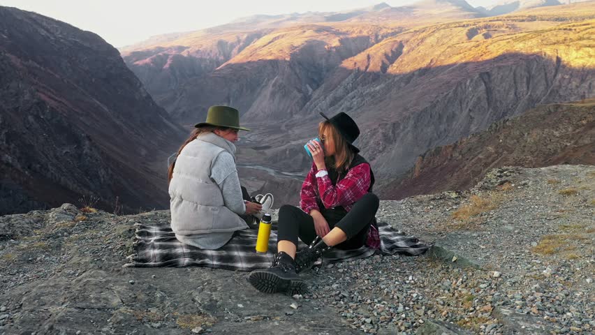 Two traveler female friends sitting on the edge of a sheer cliff above a deep cliff. Girld smilling and lookinh at the camera. Females watching the beauty of nature mountain forest and sunset on