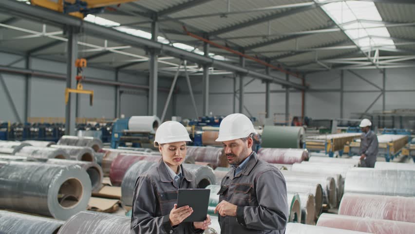 Caucasian colleagues working together on tablet device while standing in warehouse. Hardworking engineers talking about improving produce goods in factory. Workers servicing equipment in background | Shutterstock HD Video #1111440327