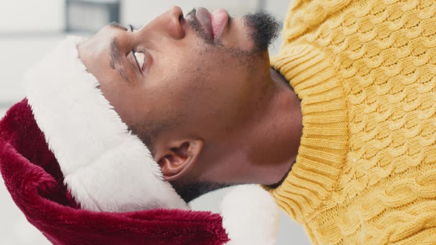 Portrait of attractive African American man wearing Santa Claus hat while positively glancing at camera. Bearded male cheerfully smiling during celebration of Christmas or New Year. Vertical view.