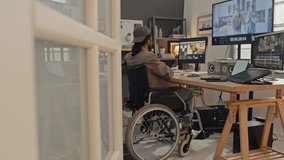 Full side shot of young man with disability sitting in wheelchair at desk at home, putting on headphones and proceeding to work on video clips in professional editing software