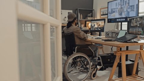Full side shot of young man with disability sitting in wheelchair at desk at home, putting on headphones and proceeding to work on video clips in professional editing software Video de stock