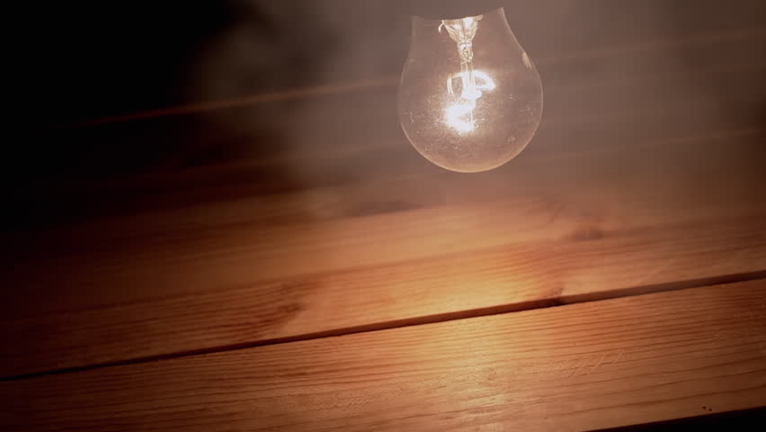 One Swinging Glowing Incandescent Light Bulb in Thick Smoke on Wooden Background. Dusty switched-on old Edison light bulb shines a bright yellow light over a table in a dark, smoky room. Texture. | Shutterstock HD Video #1111449389