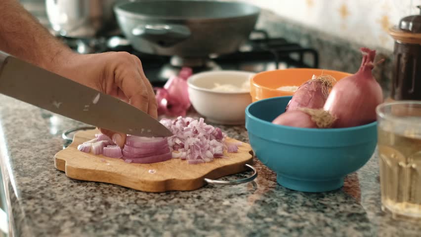 Close up of male hands cut fresh red onion on wooden cutting board on background of vegetables and greens in kitchen. Chop. Cooking food. Preparing lunch at home with healthy raw ingredients.Chopping