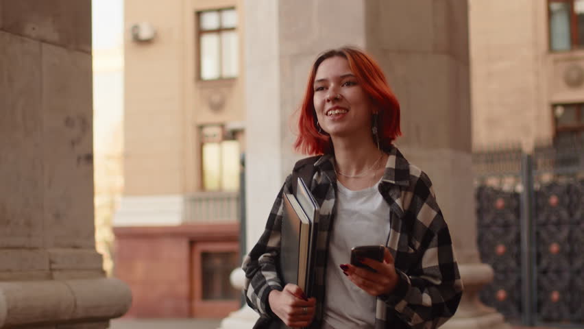 Young attractive girl with red hair wearing casual clothes walking aside the building with a laptop, smiling and reading messages on a smartphone