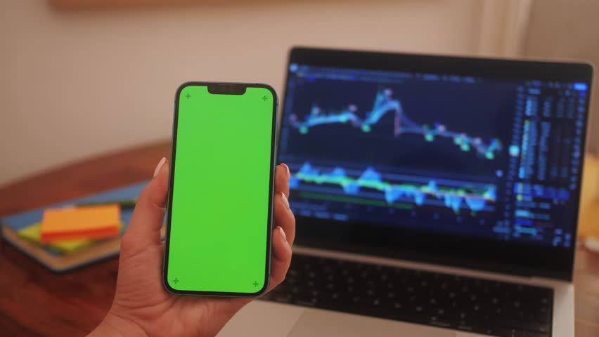 Trader holds smartphone in hands with green screen on chart of movement of cryptocurrency on screen of monitor laptop background. Stock market data statistics using mobile app. Royalty-Free Stock Footage #1111472743
