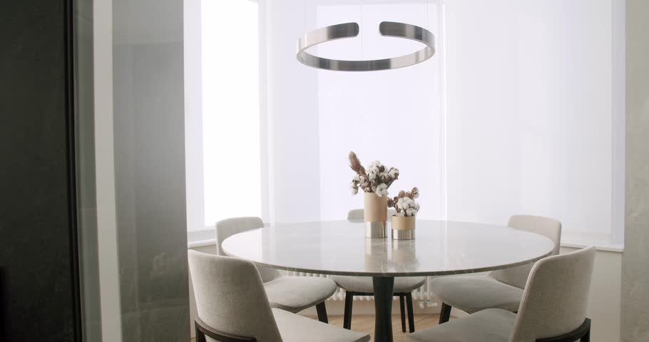 Dining round table with marble surface, comfort chairs and vase with cotton branches behind glass door at stylish apartment. Detail in interior. Elegant furniture in modern house. Decoration idea