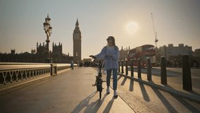 Stylish young tourist woman walking with bicycle on Westminster Bridge London with Big Ben view at sunset. Concept of eco-friendly transportation and healthy lifestyle in London - Powered by Shutterstock - Get 15% off with code: PIKWIZARD15