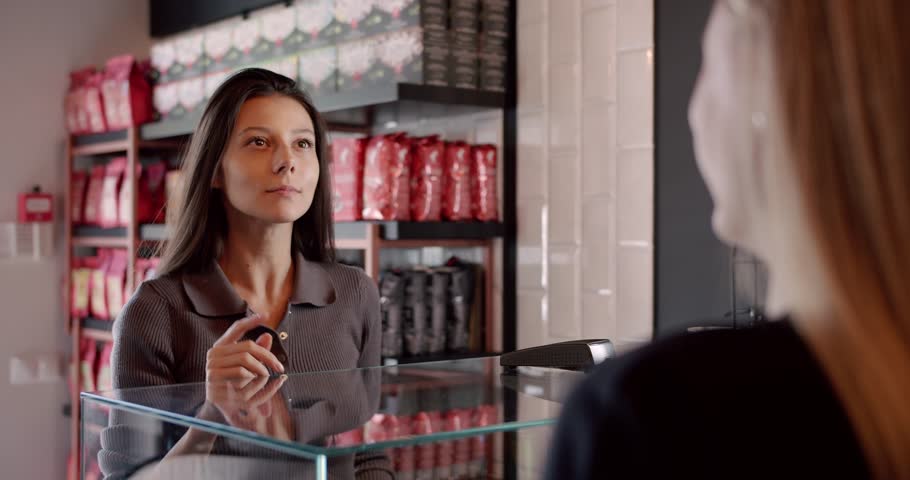 Thoughtful young female customer with long dark hair squinting and smiling while choosing coffee and communicating with crop unrecognizable barista in modern cafe.