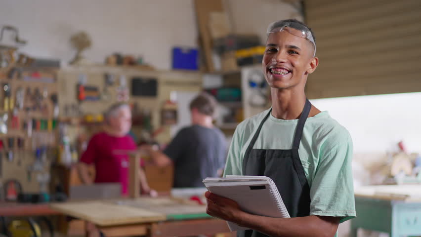 Portrait of a young black Brazilian worker holding notepad and uniform inside carpentry workshop. Job occupation concept