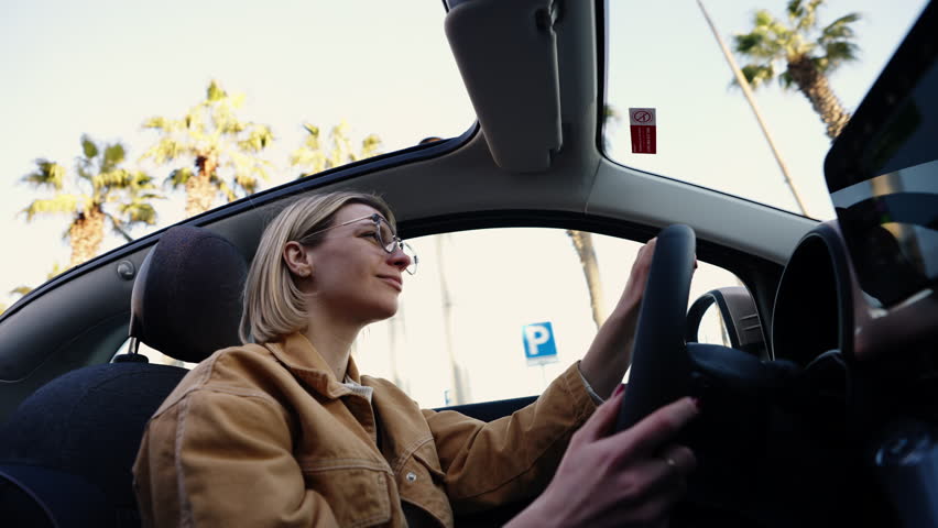 Blonde woman smiling while driving car, sunroof open with palm trees in the background, embodying joy and the essence of a carefree lifestyle