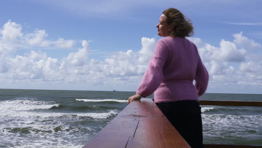 A woman in a pink jacket stands on the observation deck, looking at the sea. Her hair flutters in the wind, she smiles, relaxes.