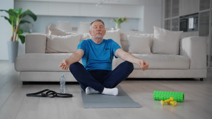 Relaxed senior man doing yoga meditation sitting in lotus pose on mat in living room at home. Elderly gray-haired bearded man making physical exercises. Yoga, sport lifestyle, exercising concept.