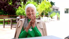 Joyful elderly lady applauding with a wide smile, seated in a nursing home's garden, captured in 4K.