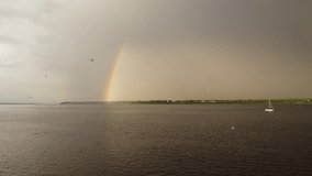 Video of a small sailboat on the water after the storm with stormy clouds and part of a rainbow, with birds flying around