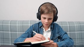 Homeschooling, distance learning online by video call, teen boy sitting at workplace in living room and uses a webcam to study in self-isolation at home, the boy in headphones writes in a notebook.
