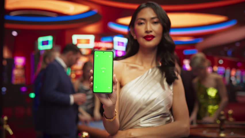Advertising Template with an Asian Female Showing a Smartphone Device with a Green Screen Mock Up Display with Trackers. Professional Beautiful Model in a Casino with People Gambling in the Background | Shutterstock HD Video #1111517955