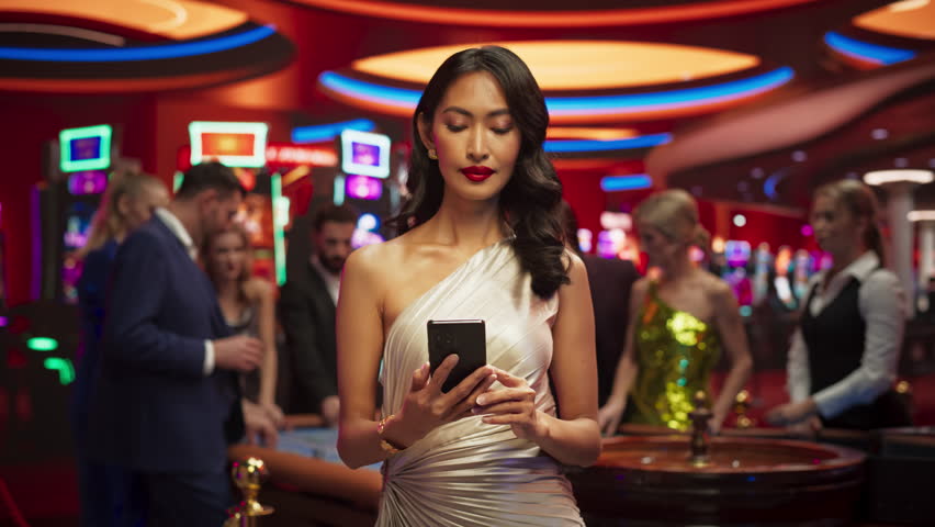 Modern Casino Setting: Gorgeous Asian Brunette Woman Posing with a Mobile Phone with a Green Screen Placeholder Space for Advertising or Commercial Materials for a Gambling Industry Royalty-Free Stock Footage #1111517957