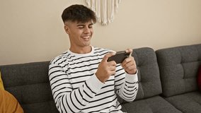 Happy young hispanic male gamer celebrating victory while playing video game, smiling man enjoys winning on smartphone in the comfort of living room sofa, at-home fun tech celebration