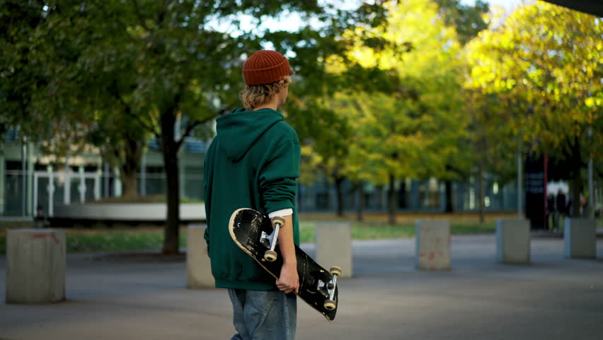 Trendy cool man holding skate board and looking away while walking at the urban park | Shutterstock HD Video #1111524411