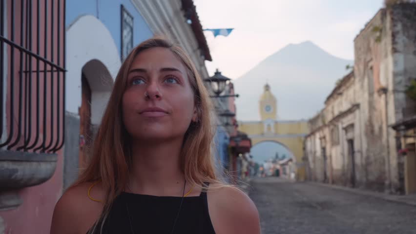 Young beautiful blonde girl with blue eyes smiles at camera, traveling in colonial city of Antigua Guatemala with cobblestone streets and yellow Santa Catalina clock tower visible in the back.