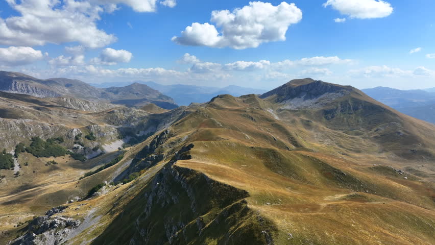 Flight over beautiful mountain peaks covered with grass. Mountain from above on a sunny autumn day. High rocks of mountain peaks that merge with mountain meadows with puffy clouds in the background.