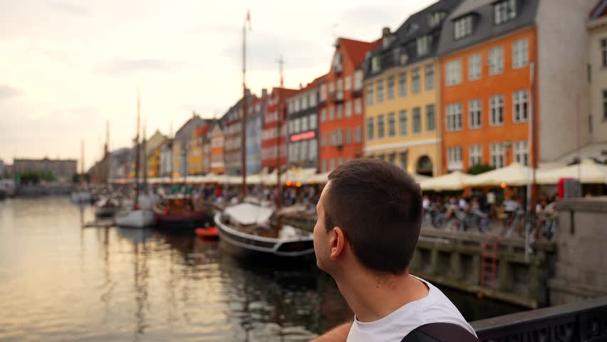 Man enjoying sunset from bridge in Nyhavn with view of boats and colorful townhouses. Tilt-down