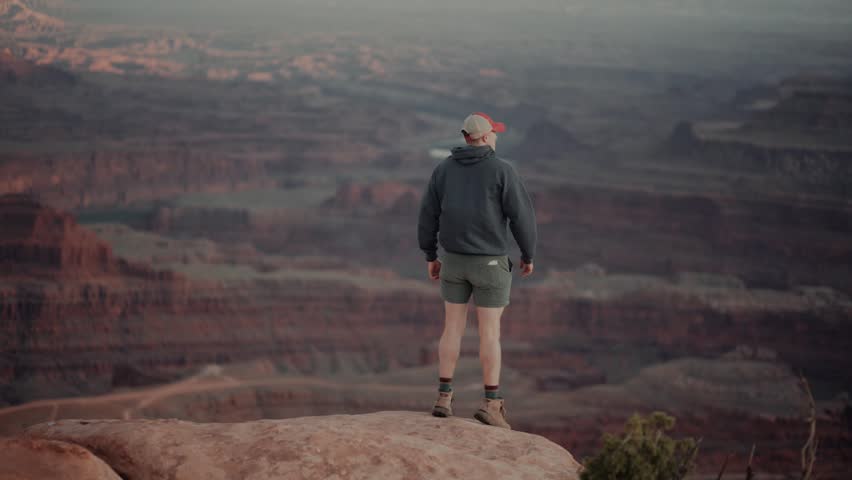 Male hiker walks to the edge of a cliff and admires stunning canyon views during sunset