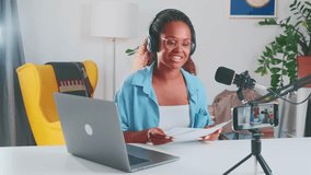 Young successful popular African American woman financial blogger records video on smartphone camera and applauds demonstrating documents with business diagrams sits at table in spacious apartment.
