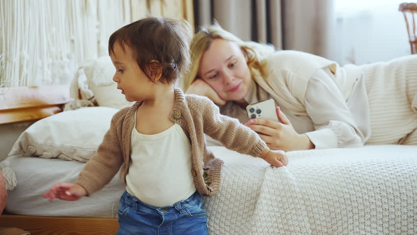 Mother using phone to play cartoon movie to make little baby happy on bed.