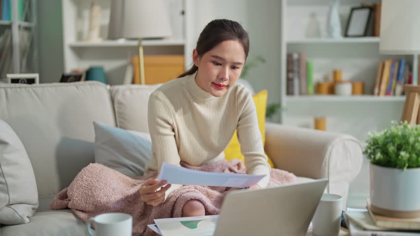 Woman going through bills, looking worried. Young adult asian woman female reading her bill papers and getting headache from stress Young asian woman at home paying online bills