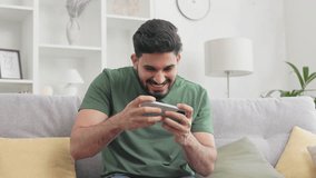 Bearded indian man emotionally playing video games on smartphone while sitting on grey couch. Irritated young guy displaying dissatisfaction while experiencing loss at modern living room.