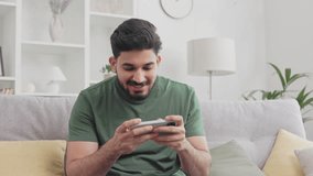 Excited bearded guy in casual wear sitting on couch and using modern smartphone for playing video games. Indian male gamer relaxing on comfy couch and dissatisfied with result while losing.