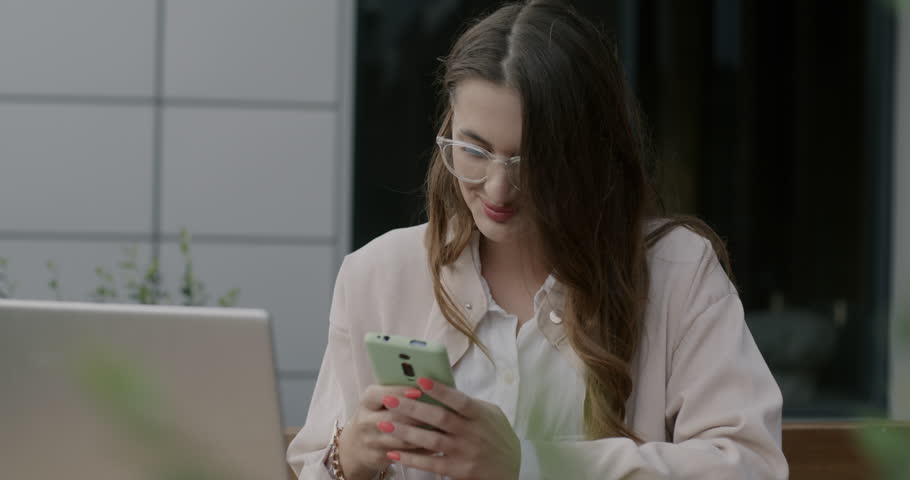 Joyful young woman using smartphone and laughing sitting in street cafe with laptop alone. Modern technology and youth lifestyle concept.