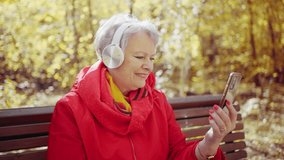 An elderly gray-haired woman in headphones and a red jacket uses a smartphone for video communication