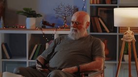 An elderly man in good health and positive mood sits in the room in an armchair, uses a smartphone and video call conversation. A human waves hand as greeting sign, smiles, talks animatedly and