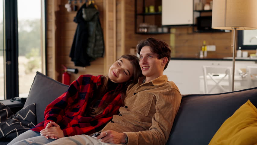 Smiling couple in love a guy and a girl are sitting together on the sofa and watching comedy film in a cozy wooden house