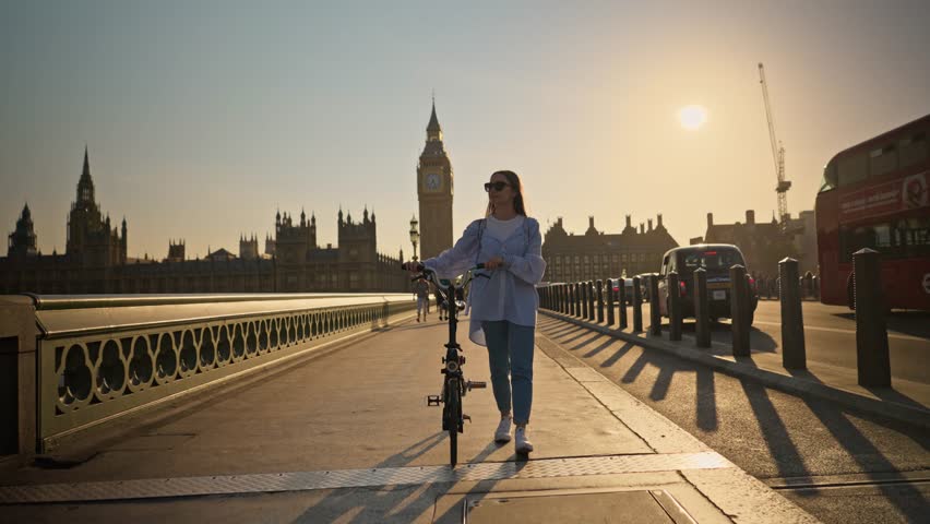 Stylish young tourist woman walking with bicycle on Westminster Bridge London with Big Ben view at sunset. Concept of eco-friendly transportation and healthy lifestyle in London Royalty-Free Stock Footage #1111571801