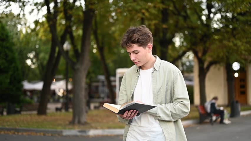 A young guy walks through the park and reads a paper book