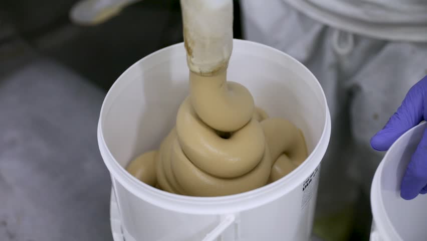 Food industry. Ice cream production process. Closeup view of caramel ice cream coming out from a pipe and into a commercial plastic container. The factory worker guides the process.