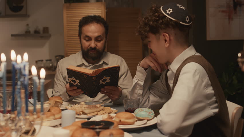 Medium shot of devoted Jewish father in kippah sitting at Hanukkah dinner with family and reading story from holy Torah to teenage son in skullcap and daughter, children listening attentively Royalty-Free Stock Footage #1111580913