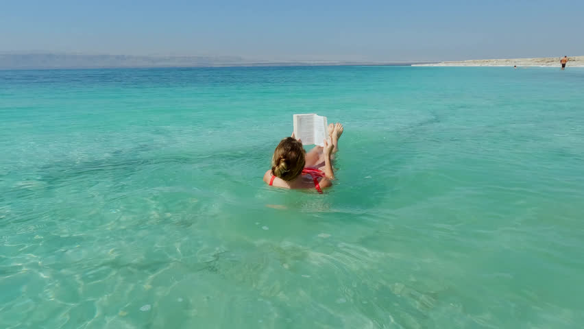 Beautiful Young Woman Floats in the Dead Sea in Red Swimsuit while Reading Book